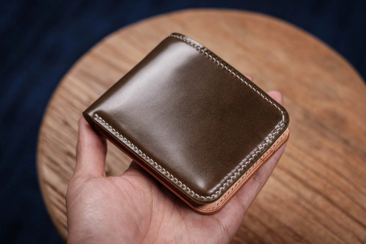 A bifold wallet featuring a smooth, olive-green exterior with a glossy finish and light-colored stitching along the edges. The wallet has a natural, light tan leather interior visible at the bottom edge, showcasing a layered construction.