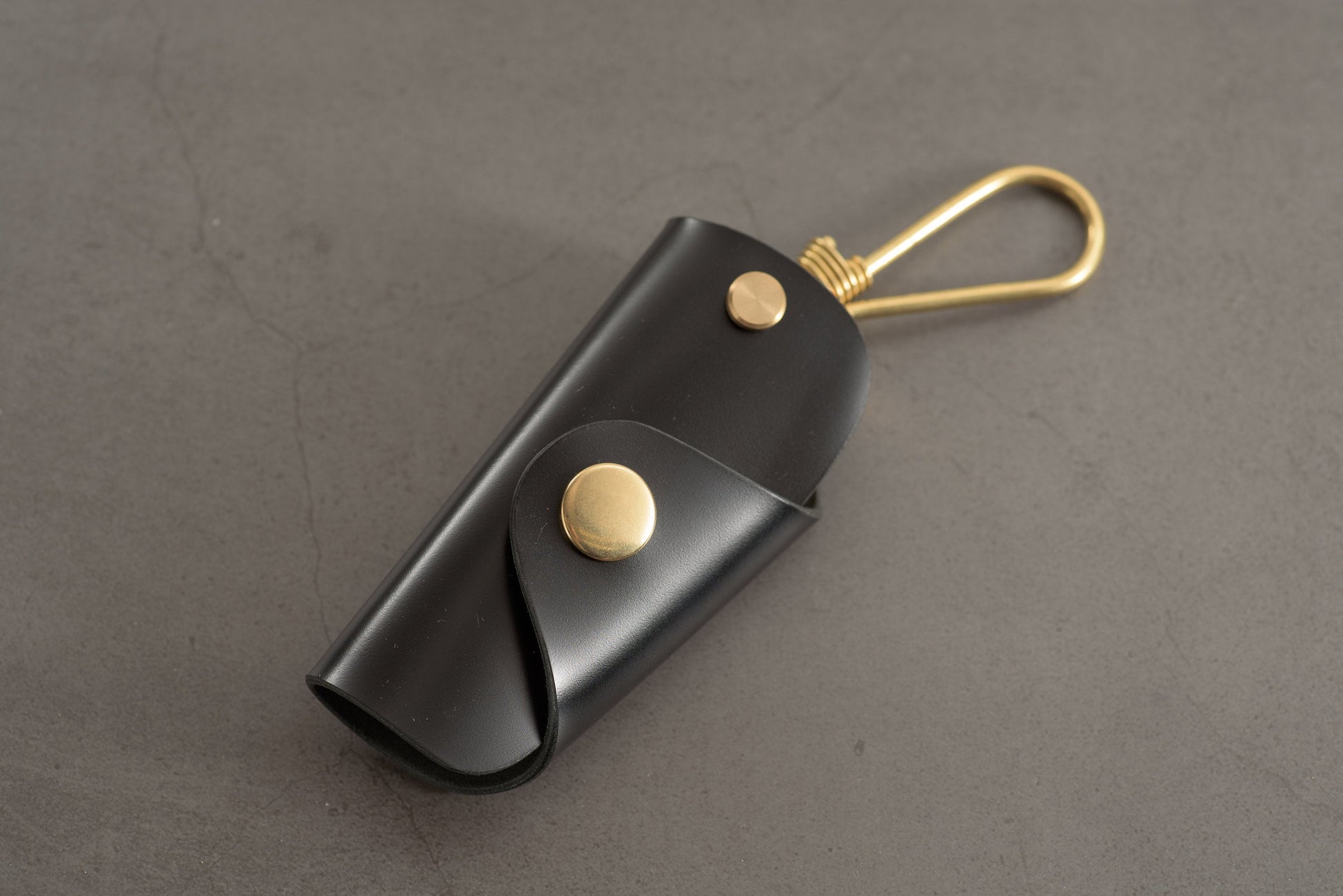 A black Shell Cordovan leather key case with brass hardware and a removable brass wire hook.