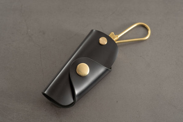 A black Shell Cordovan leather key case with brass hardware and a removable brass wire hook.