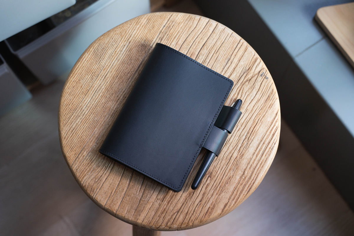 A black leather notebook with a pen holder and a black pen. The notebook is sitting on a wooden surface.