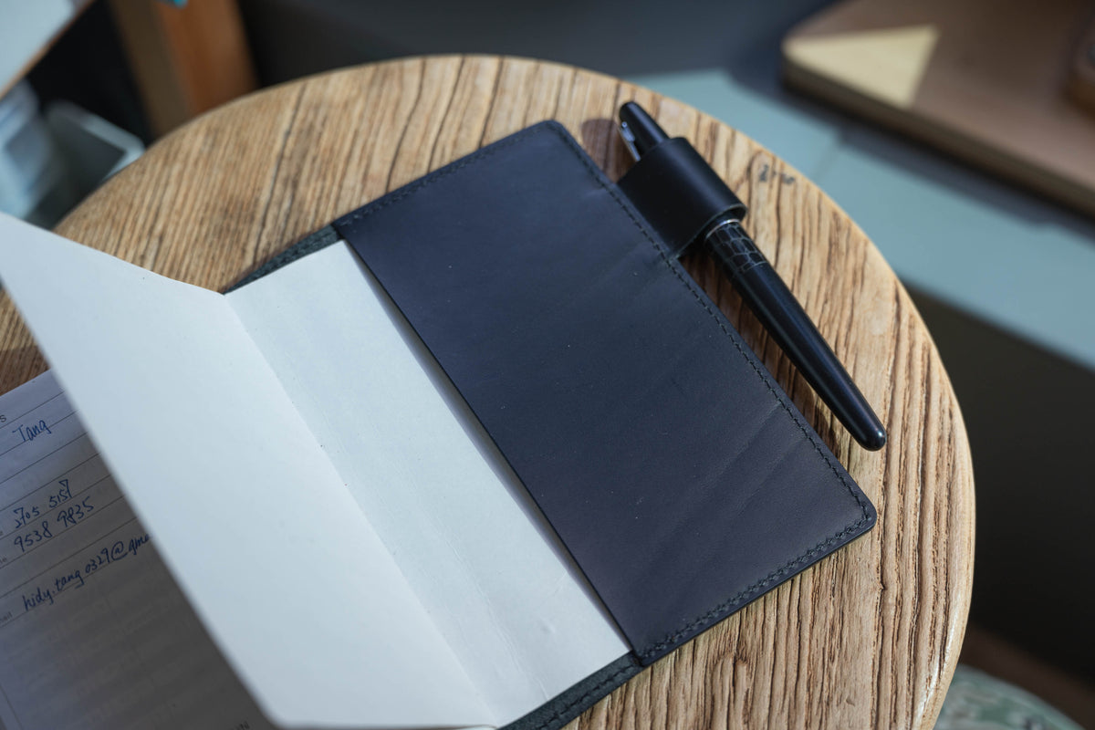 A black leather notebook cover display with black pen and notebook inside.