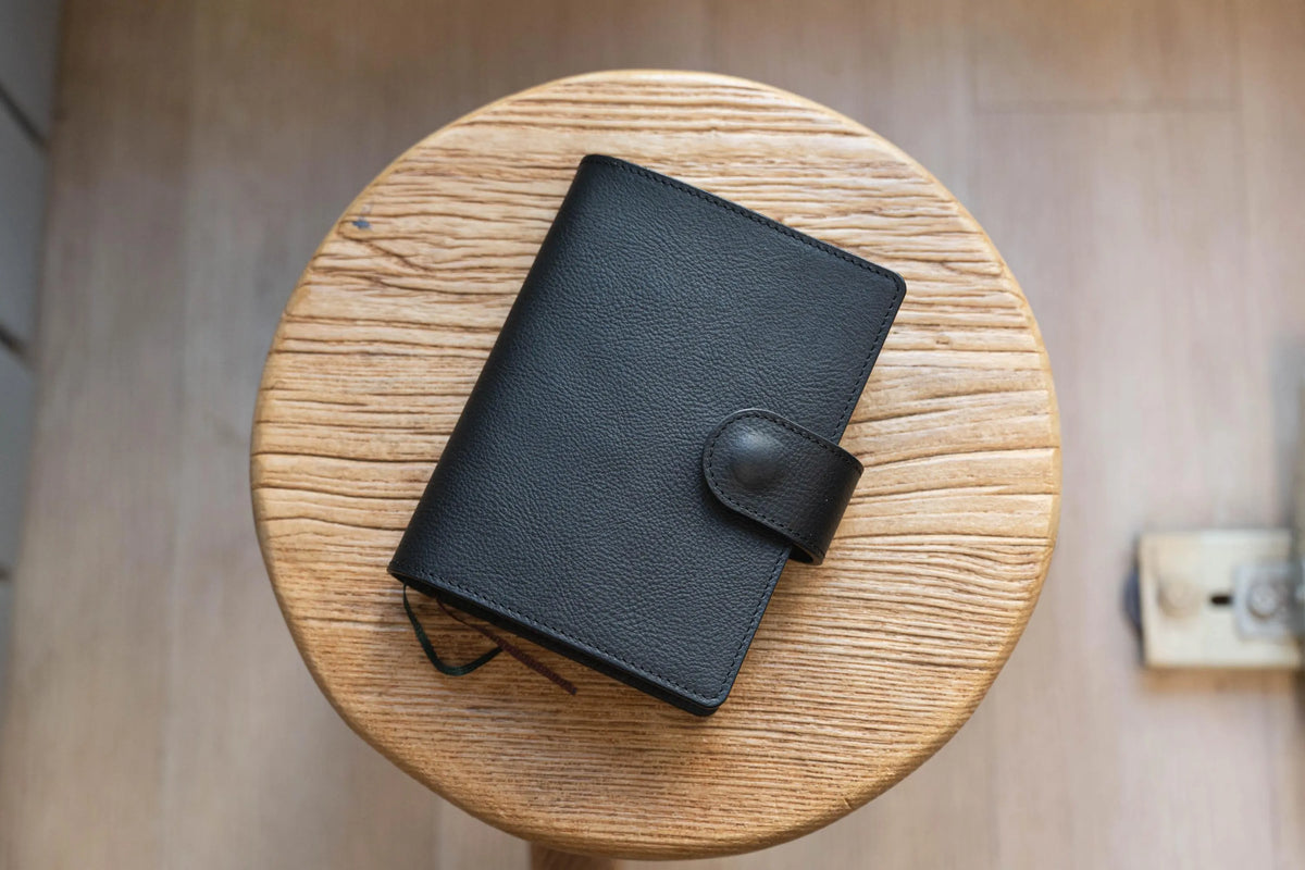 A black, rectangular, textured notebook cover with a rounded flap closure secured by a matching black button. 