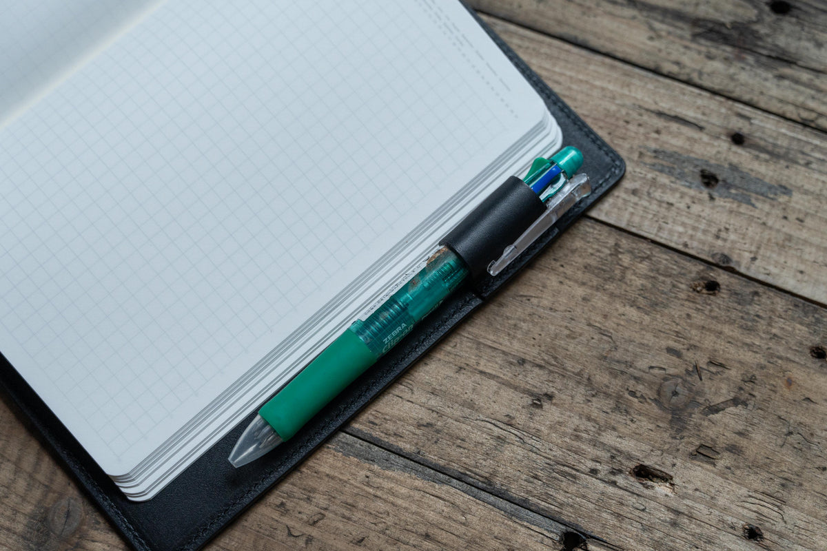 Close up for the pen loop of a black notebook cover. A green pen insert into the pen loop.