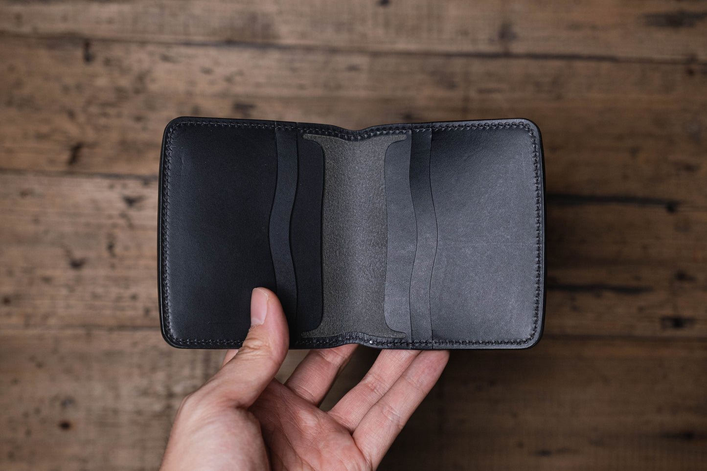 A black leather bifold wallet featuring a smooth exterior finish and an interior with multiple card slots. The wallet is constructed with visible tonal stitching along the edges and includes two vertical card pockets on each side.