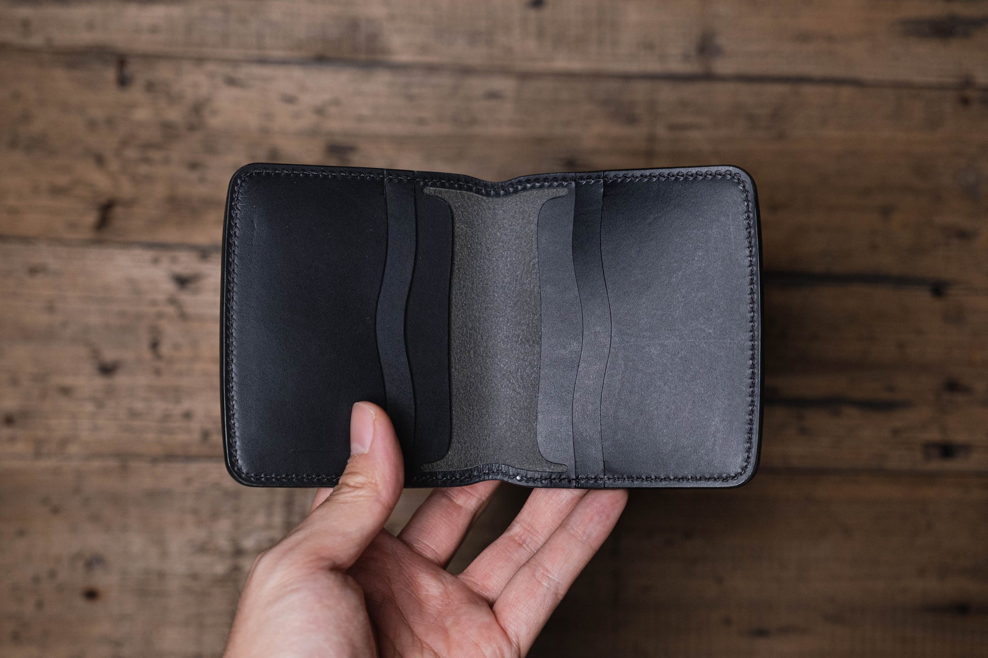A black leather bifold wallet featuring a smooth exterior finish and an interior with multiple card slots. The wallet is constructed with visible tonal stitching along the edges and includes two vertical card pockets on each side.
