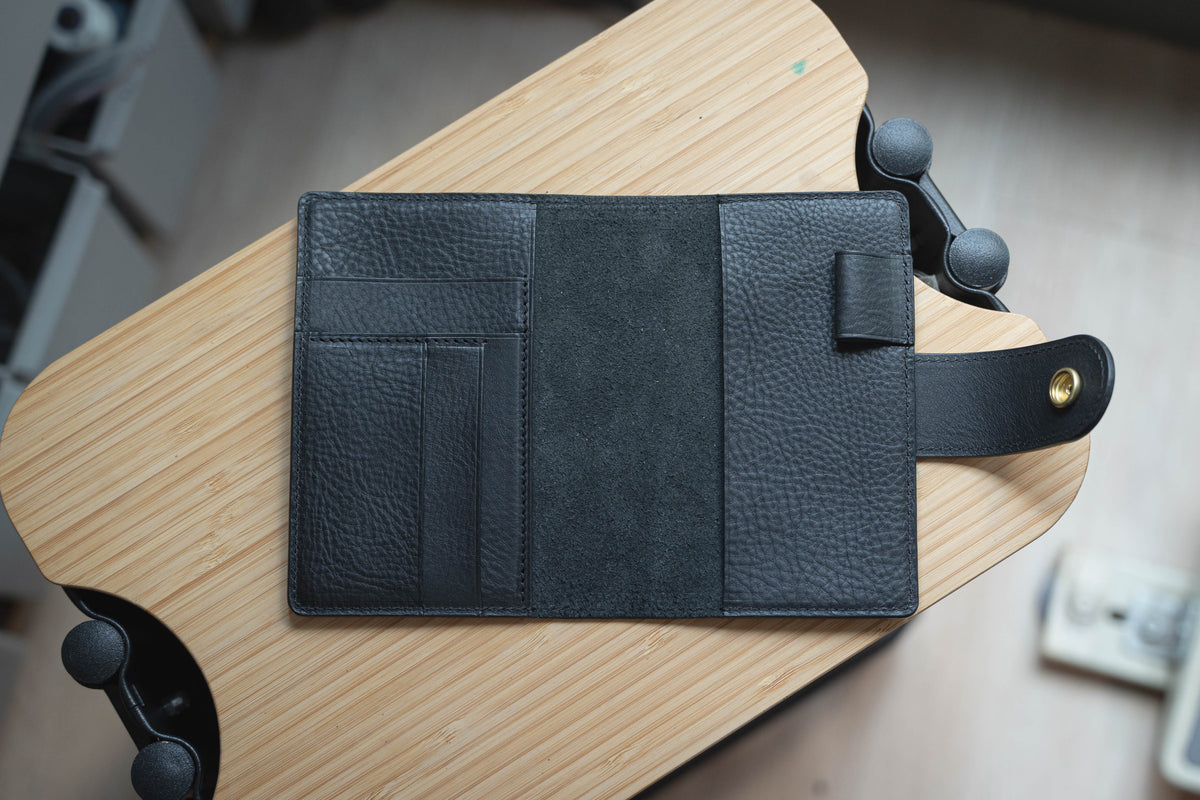 A black leather notebook cover with a flap closure and a pocket on the front cover. The notebook cover is open.