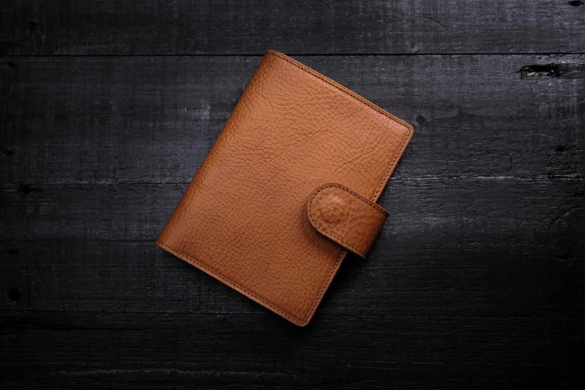 A brown leather notebook cover with a pebbled texture and visible stitching along the edges. The cover features a small, rounded flap closure on the right side that secures the front panel to the main body of the notebook.