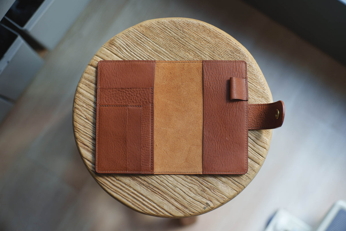  A brown leather notebook cover with a flap closure and a strap with a brass button. The cover has a pocket on the front and a flap on the back.