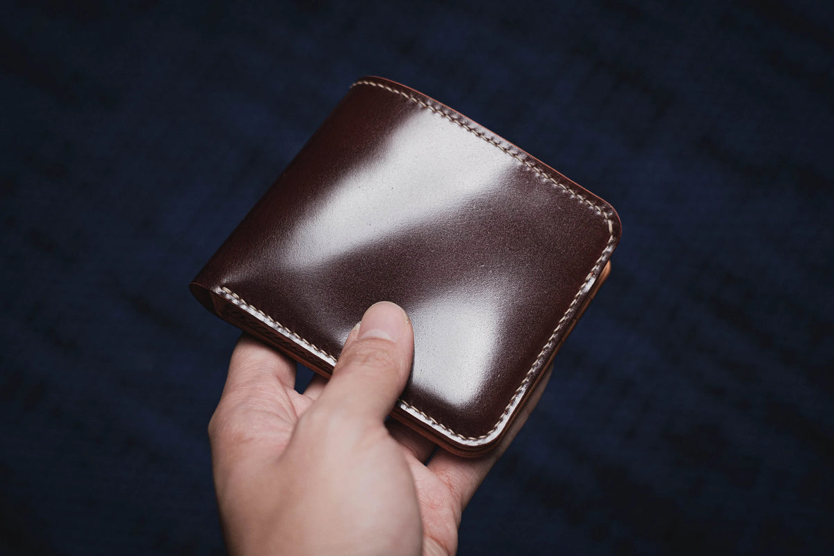 A hand holding a burgundy leather bifold wallet with white contrast stitching against a dark textured background.