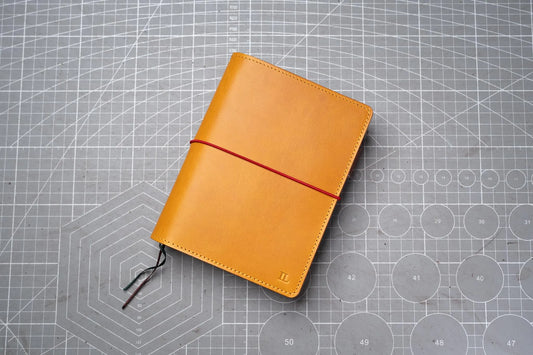 A mustard yellow leather notebook cover featuring a smooth finish, stitched edges, and a thin red elastic band wrapped horizontally around the center. 