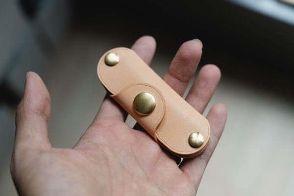 A hand holding a natural leather key holder with brass snaps and screws.