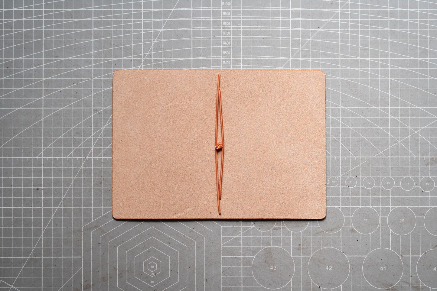 A square tan leather folio or notebook cover with a vertical center seam, laid flat on a gray cutting mat with grid lines.