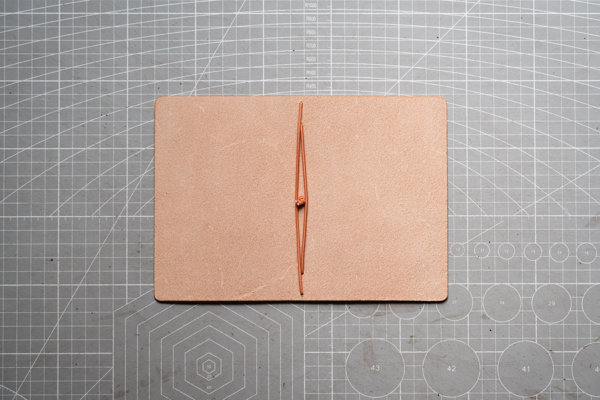 A square tan leather folio or notebook cover with a vertical center seam, laid flat on a gray cutting mat with grid lines.