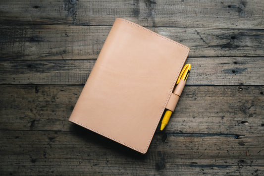 A natural leather notebook cover with visible stitching along the edges. A bright yellow pen is inserted into a matching leather pen loop attached to the side of the cover. The notebook is rectangular and features a smooth, uniform surface.