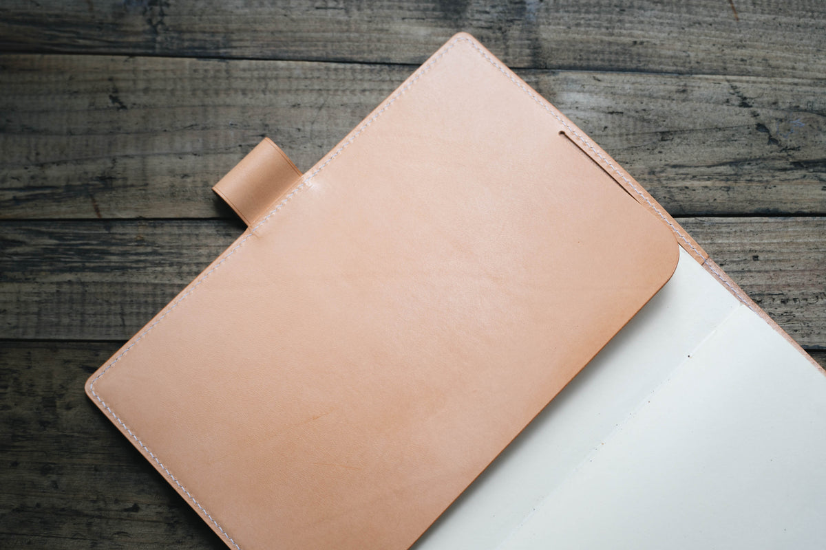 A natural leather notebook cover featuring a smooth, natural finish with visible white stitching along the edges. The cover includes a small, integrated leather pen loop on the left side and holds a stack of plain white paper pages.