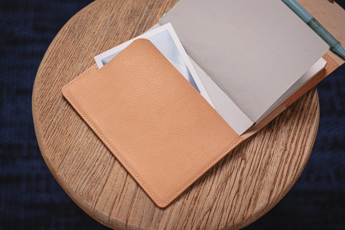 A natural leather notebook with a pocket on the front. A white piece of paper is sticking out of the pocket. The notebook is open.