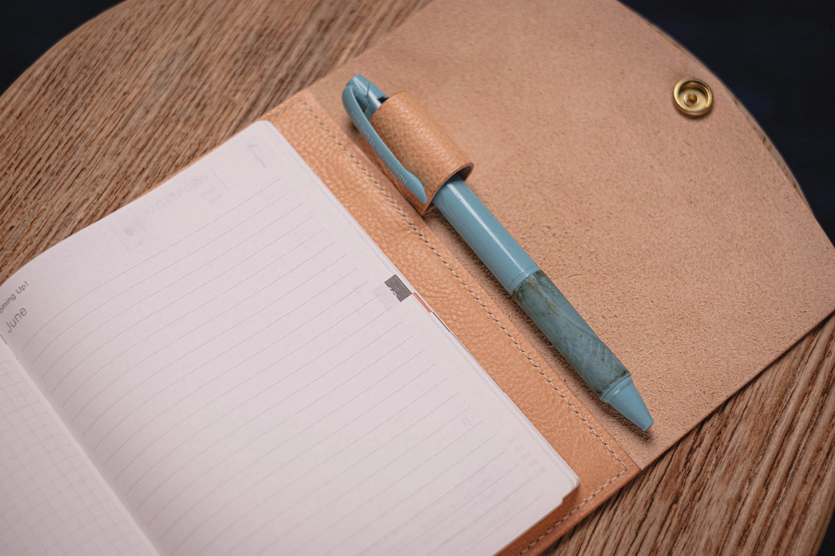 A natural leather notebook cover with a pen holder and a blue pen. The notebook is open to a blank page with lined paper.
