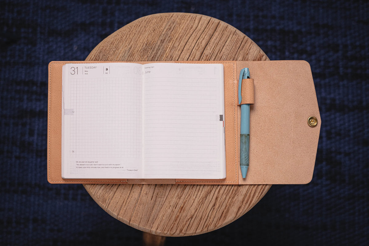 A natural leather journal cover with a blue pen tucked into the cover. The journal is open to a blank page with a grid pattern. 