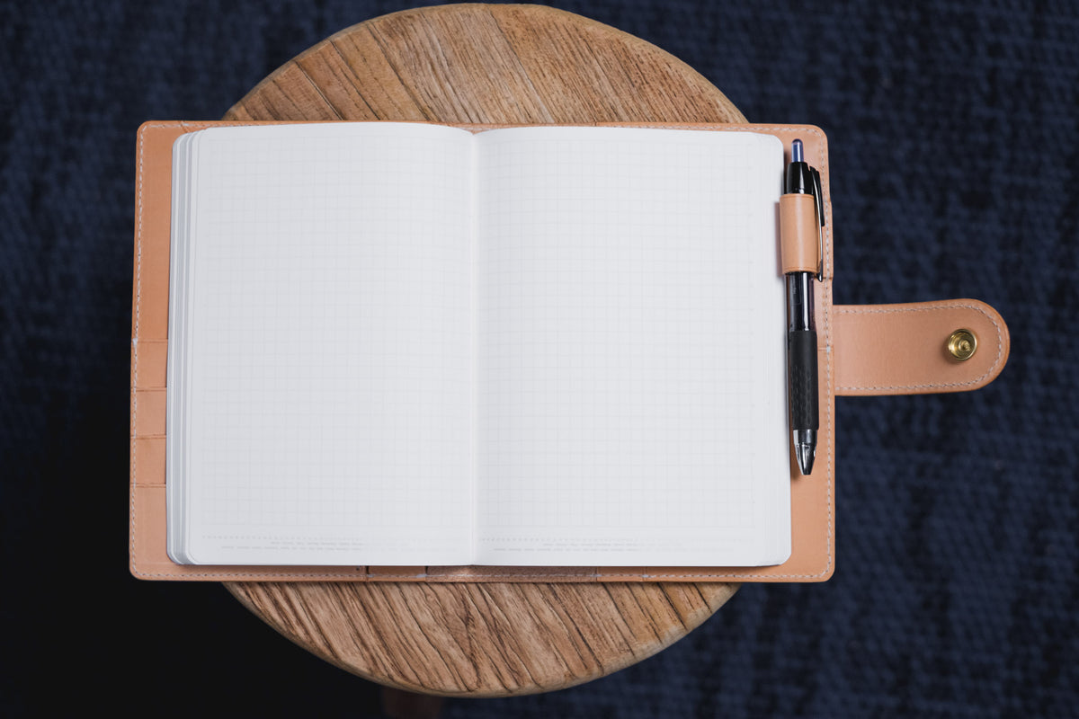 An open notebook with white grid-lined pages held within a natural leather cover. A black and silver pen is secured in a side loop on the right side of the cover, which features a small brass snap button closure on the strap.