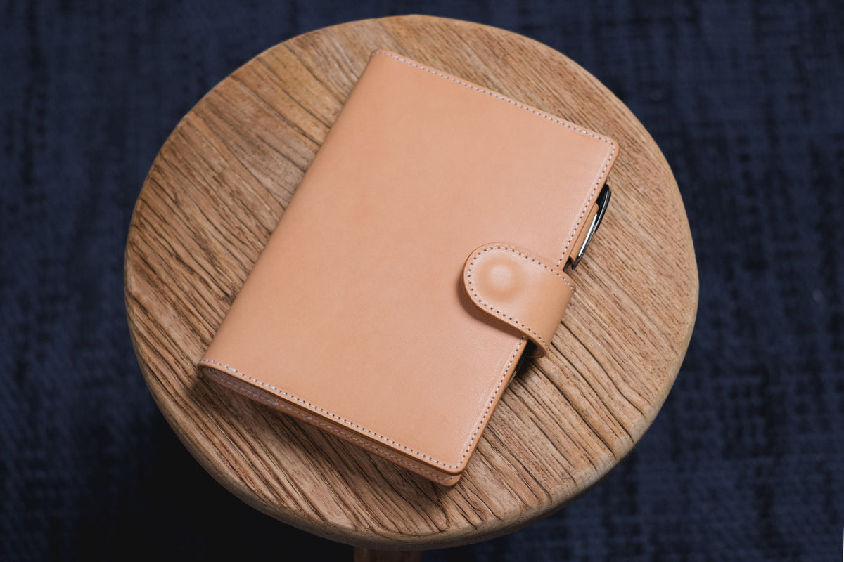 A natural leather notebook cover with a smooth, matte finish and visible stitching along the edges. The cover features a secure strap closure with a snap button and a pen holder loop attached to the side of the spine.