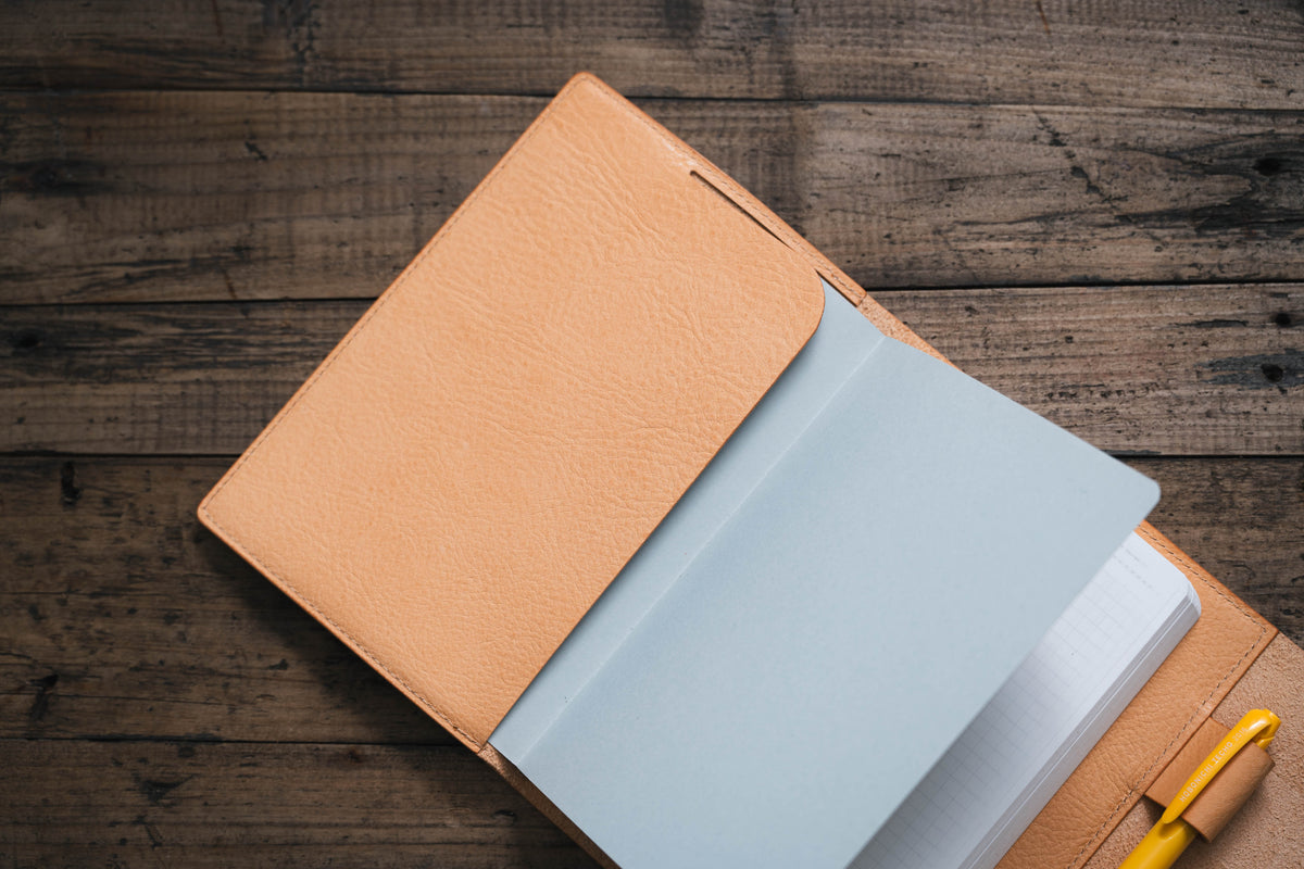 A natural leather notebook cover with a notebook inside. The notebook is open and a yellow pen is sticking out of the cover.
