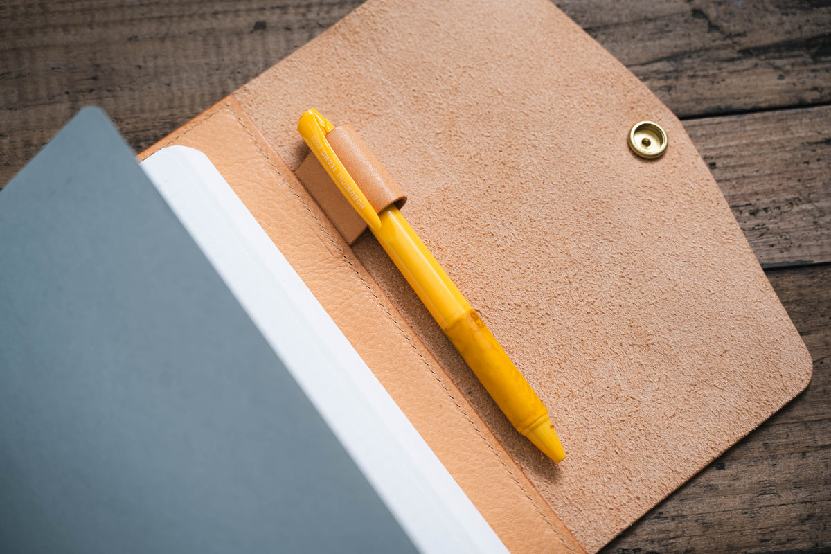 A natural leather notebook with a yellow pen tucked into a leather pen holder. The notebook is open to a blank page.