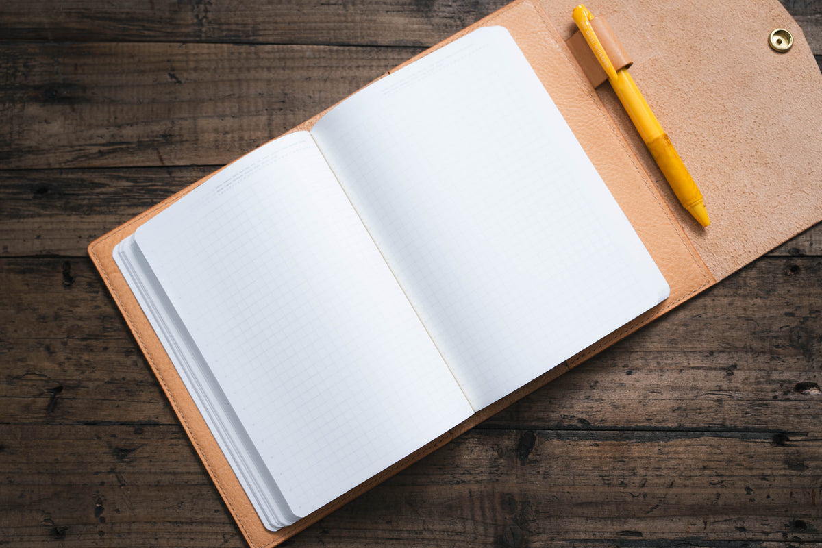 An open notebook with lined pages sits in a natural leather cover. A yellow pen is tucked into the cover.