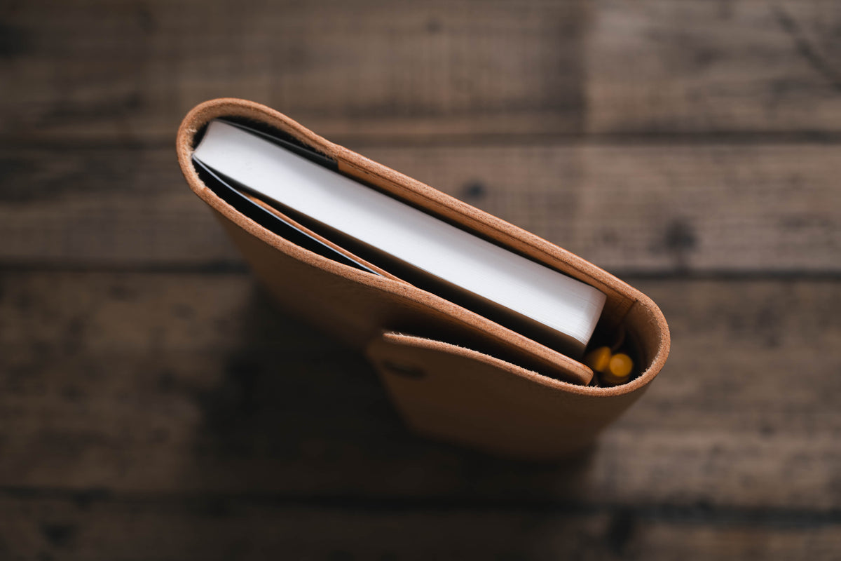 A natural leather notebook cover with a white notebook inside. The cover has a flap that closes with a snap.