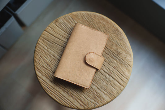 A natural leather notebook cover with a flap closure and a single button. The notebook cover is sitting on a wooden surface.