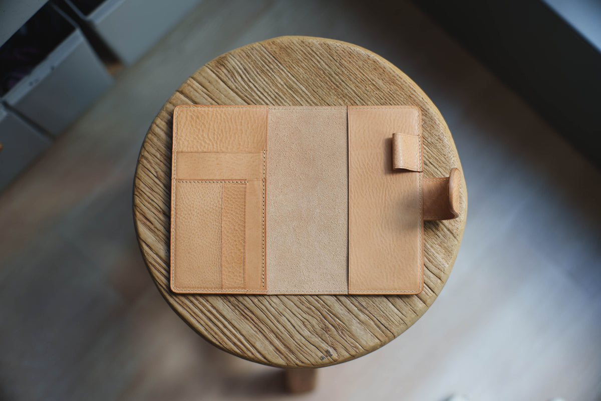 A natural leather notebook cover with a flap closure and a pen holder. The cover is made of a textured leather and has a simple design.