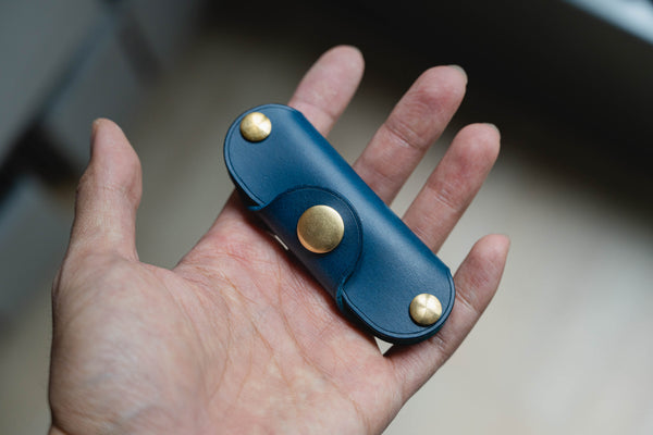 A hand holding a navy blue leather key holder with brass snaps and screws.