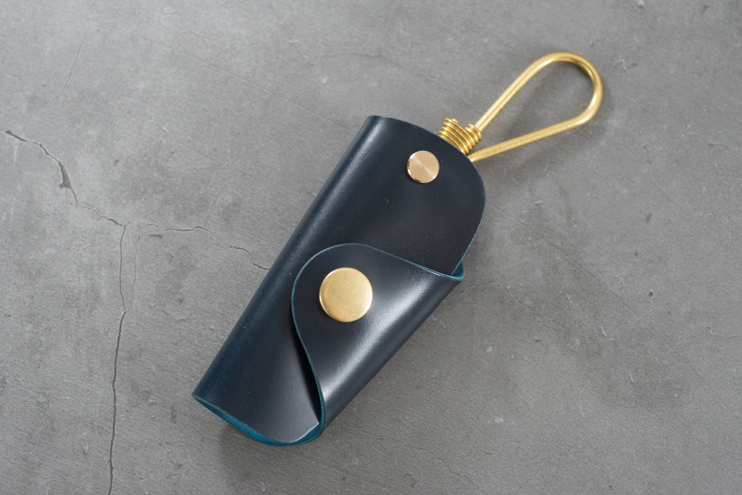 A navy blue Shell Cordovan leather key case with brass hardware and a removable brass wire hook.