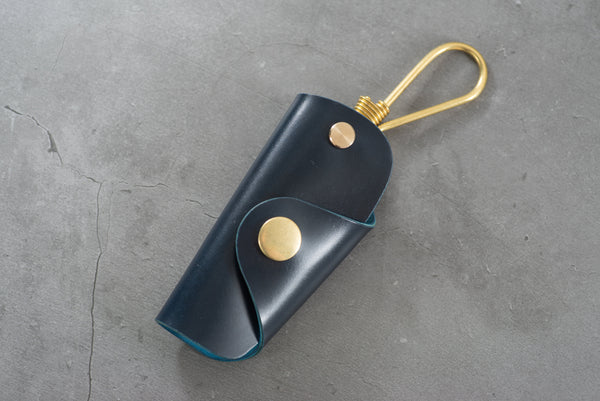A navy blue Shell Cordovan leather key case with brass hardware and a removable brass wire hook.
