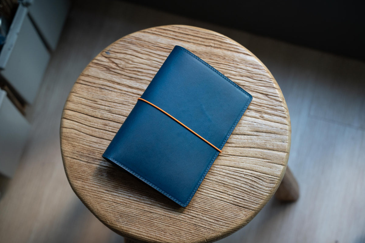 A navy blue leather notebook cover featuring a smooth, matte finish and stitched edges. A thin, vibrant orange elastic band is wrapped horizontally around the center of the cover to keep the notebook securely closed.