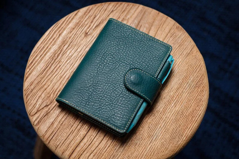 A navy blue leather wallet with a pebbled texture and a snap button closure. The wallet features light-colored stitching along the edges and a bright blue pen tucked into the side loop.