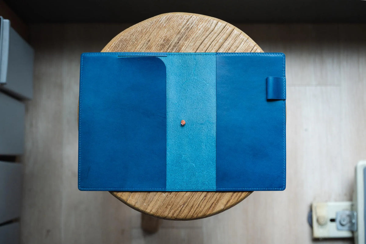 A navy blue leather notebook cover, open and lying on a wooden stool. A pen loop is visible on the right side.