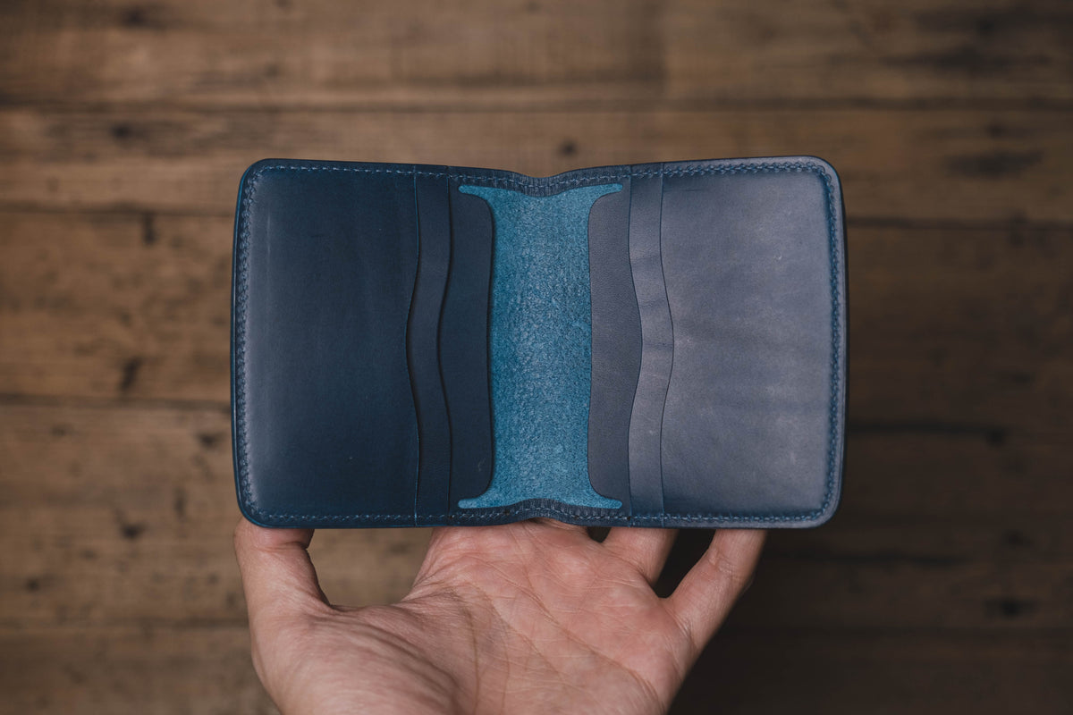 A bifold wallet crafted from smooth dark blue leather, featuring a contrasting bright navy blue interior spine and multiple card slots on both the left and right sides with curved edges and visible stitching along the perimeter.