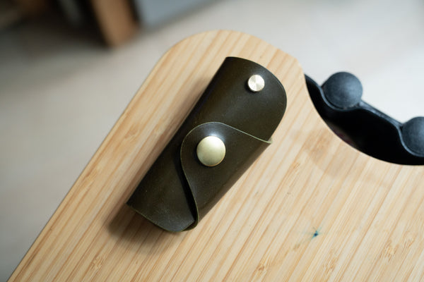 A olive green Shell Cordovan leather key case with brass hardware and a removable brass wire hook.