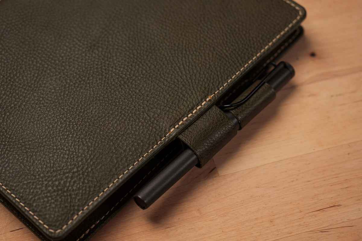 A close-up of a olive green textured leather notebook cover with contrast stitching, featuring two interlocking pen loop holding a black pen.