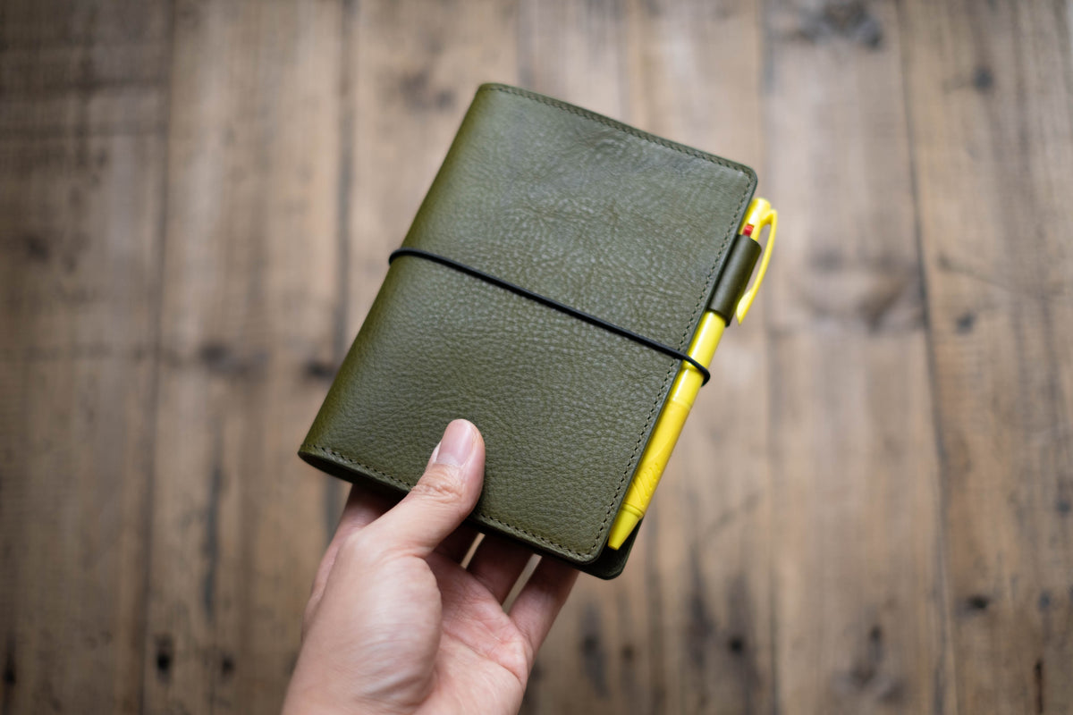 Olive green leather journal cover with a black elastic closure. A yellow pen is secured in a leather pen loop. The journal is closed and held in a hand, with a wooden background.