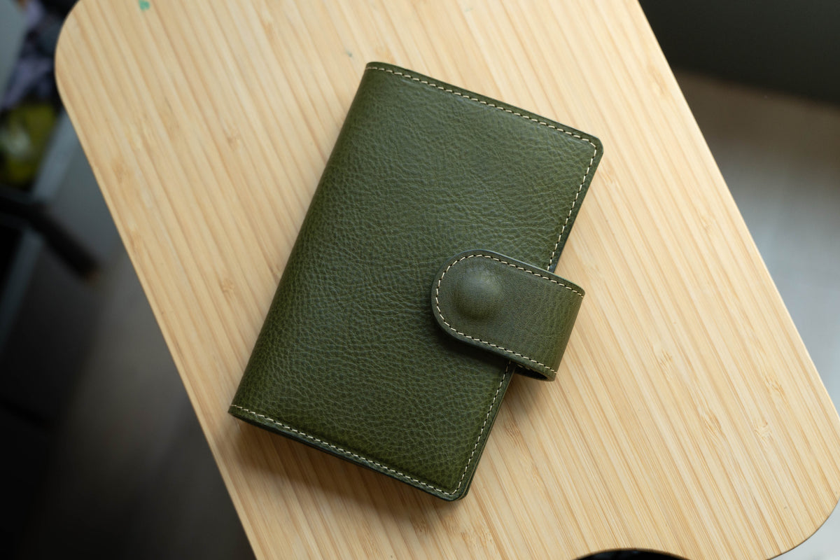 An olive green leather notebook cover with a flap closure and a stitched design. The notebook cover is closed and lying on a wooden surface.