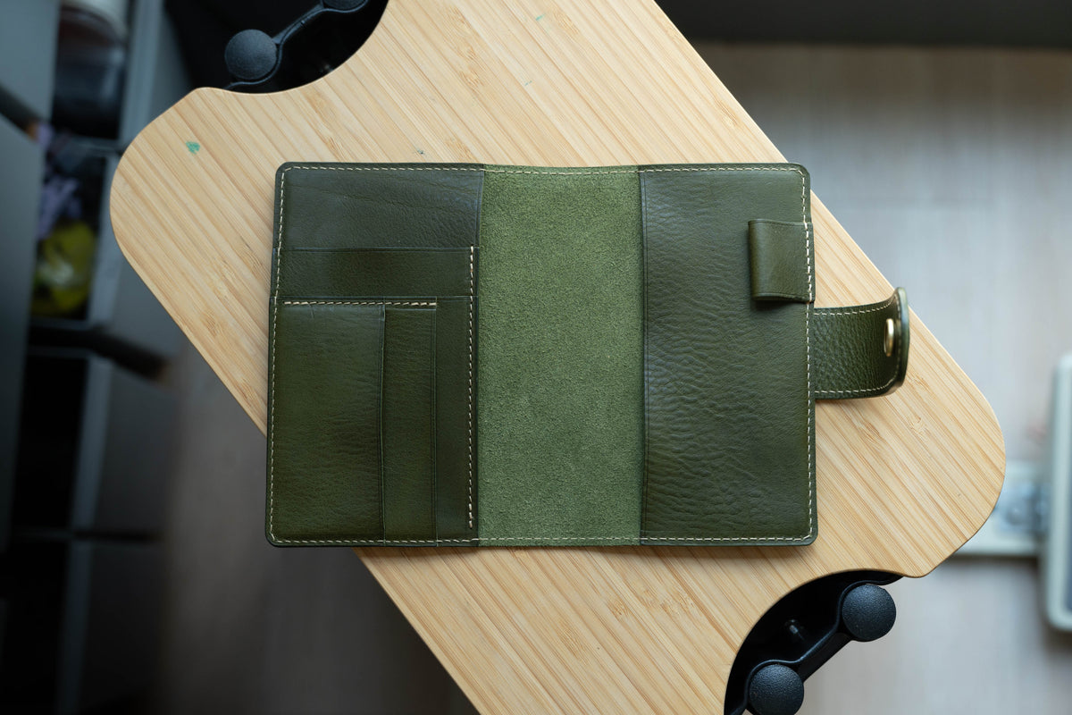 An olive green leather notebook cover with a flap closure and a pocket on the front cover. The notebook cover is open.