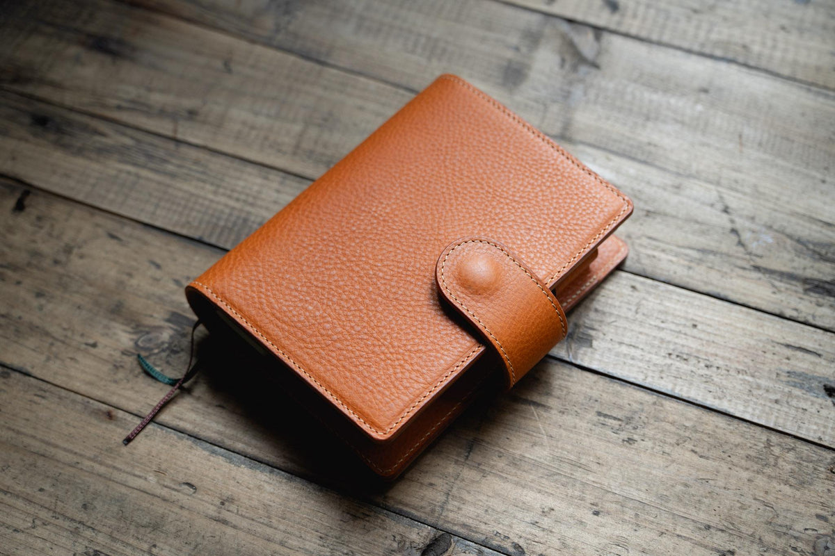 A orange brown leather planner cover with a pebbled texture and a matching strap closure featuring a circular snap button. 