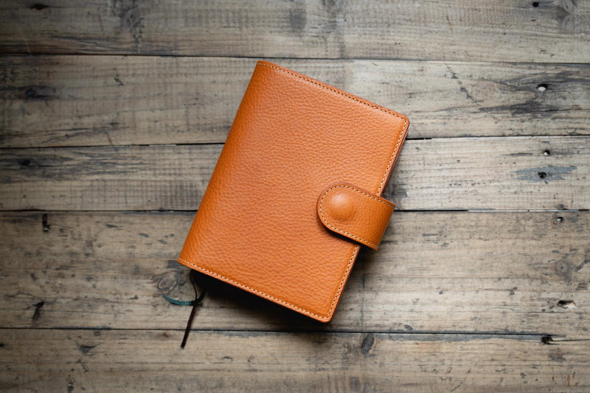 A orange brown leather planner cover with a pebbled texture and a matching strap closure featuring a circular snap button.