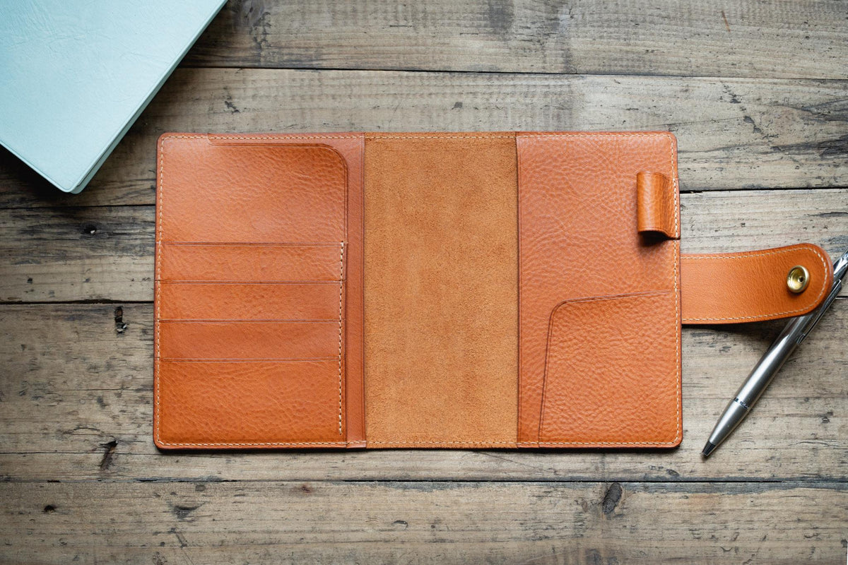 A orange brown leather notebook cover featuring a textured grain, multiple interior card slots, a central spine, a pen loop, and a side strap with a gold-toned metal snap button closure. A silver metal pen rests on the right side of the cover.