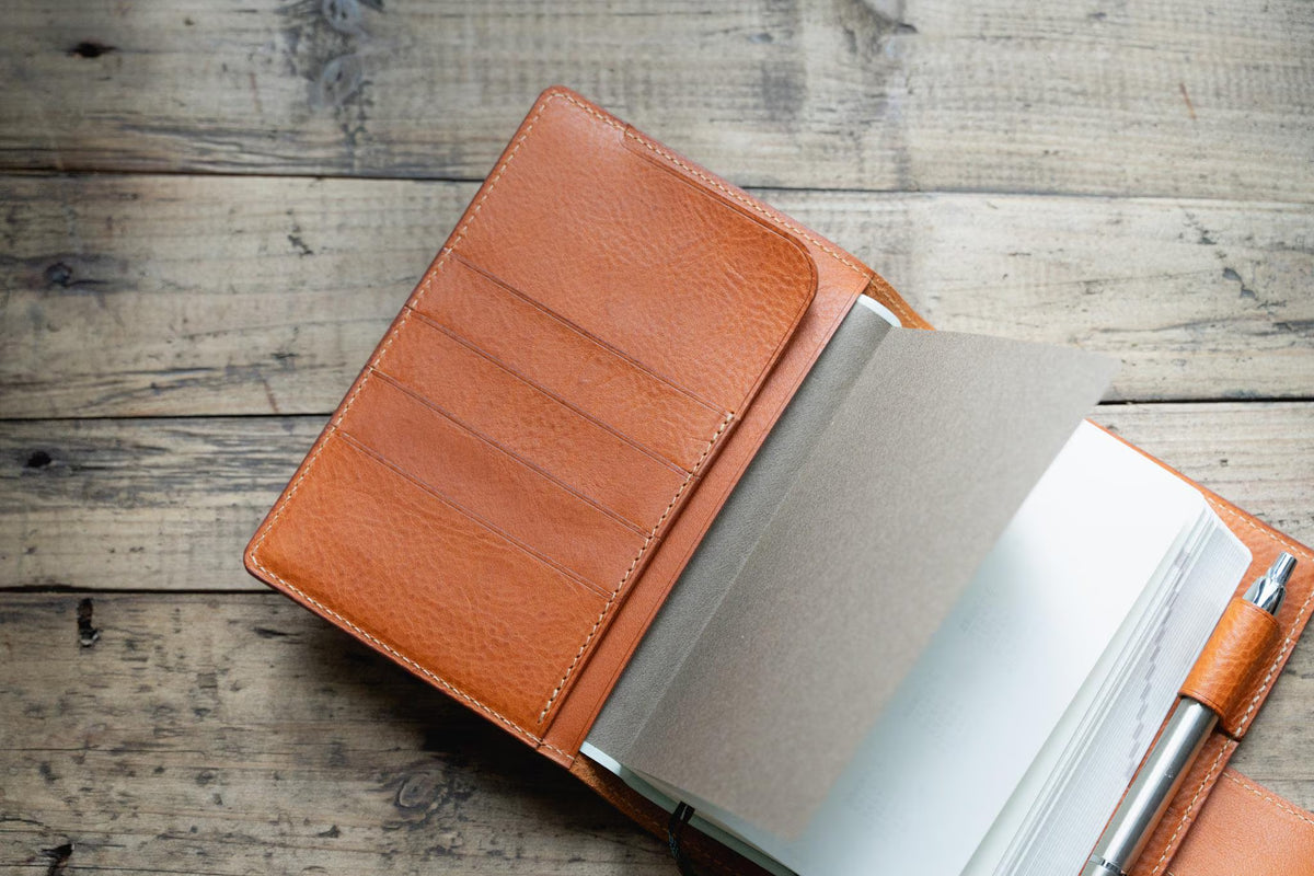 A orange brown leather notebook cover featuring a textured surface, multiple interior card slots, and a matching leather pen loop holding a silver metal pen. The notebook contains several blank white pages and a grey paper divider insert.