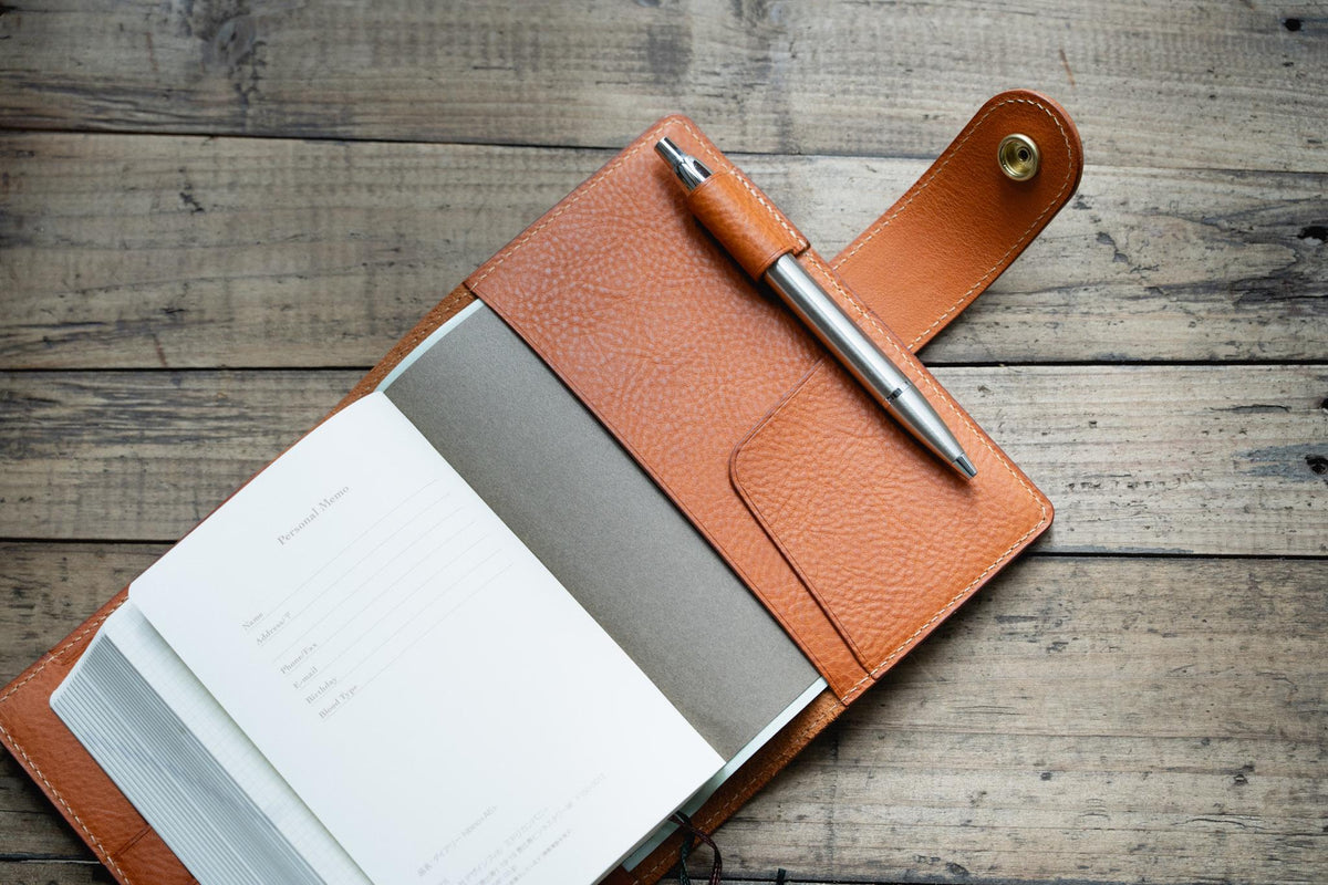 A orange brown textured leather notebook cover with a snap closure and a pen loop holding a silver metal pen. The cover contains a notebook with white pages featuring printed lines for personal information and a dark gray divider page.