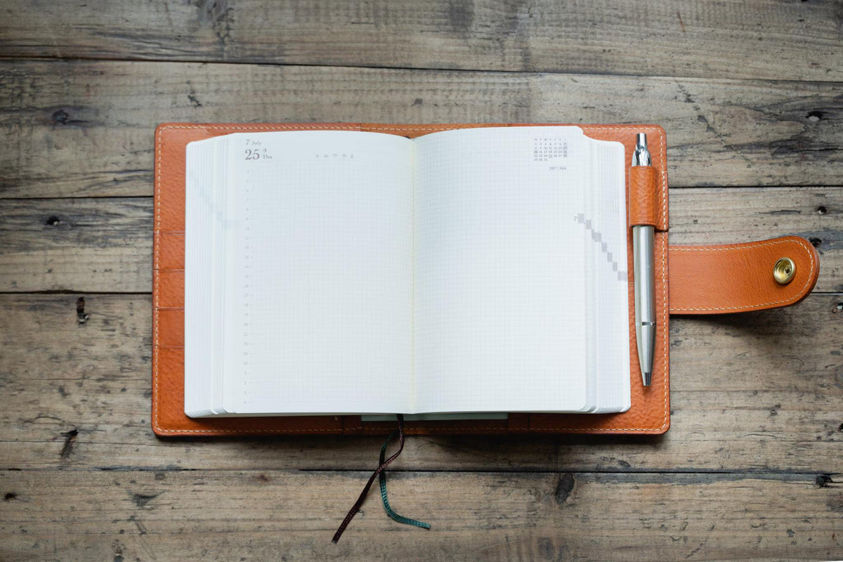 An open planner with white grid paper pages, featuring the date 25 printed on the top left corner. The planner is encased in a orange brown leather cover with a side strap and a gold-toned snap button, holding a silver metal pen in a loop.