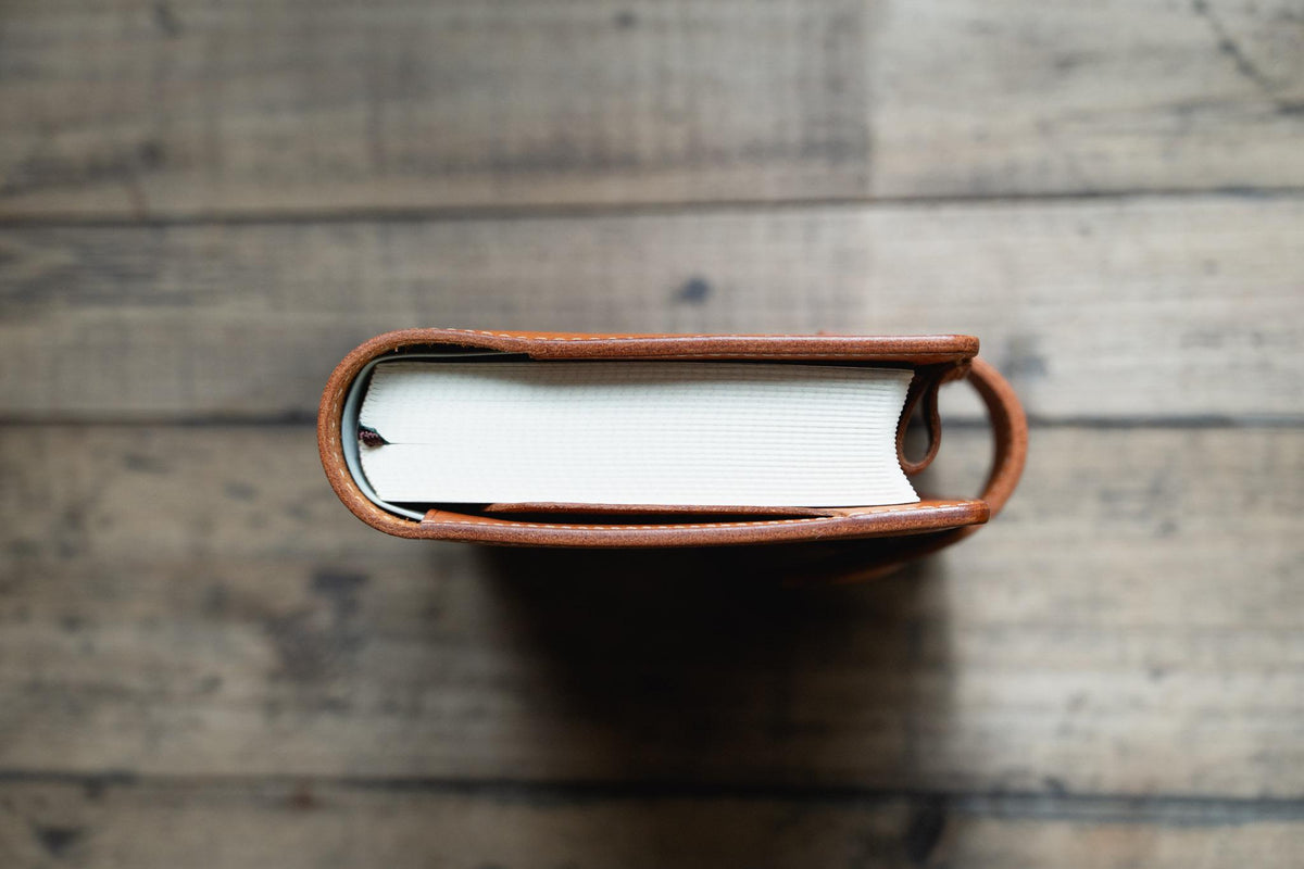 A top-down view of a thick book with white pages encased in a orange brown leather cover. The leather wrap features visible stitching along the edges and a rounded spine design that secures the pages firmly within the protective brown casing.