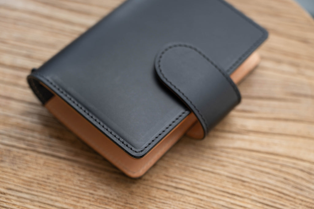 A black and natural two-tone leather traveler's notebook with card slots, displayed closed, close up of stitching and snap closure
